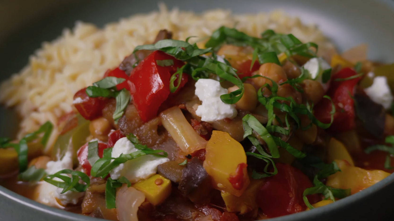Bol de ratatouille servie sur de l’orzo, avec pois chiches, poivrons rouges et jaunes, aubergine, basilic frais et fromage de chèvre émietté